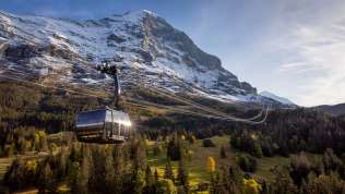 El teleférico Eiger Express en primavera, con un paisaje verde en primer plano, montañas nevadas al fondo y un cielo azul.